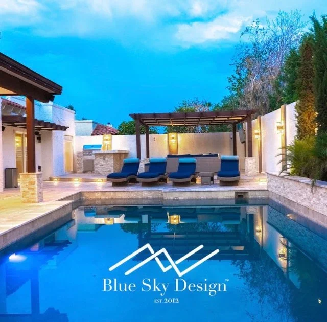 Illuminated pool with composite deck, pergola, outdoor kitchen, and lounge seating at dusk