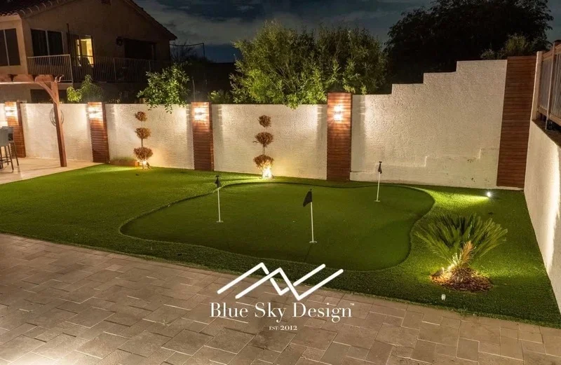 Illuminated synthetic putting green with landscape lighting, paver patio, and stucco walls
